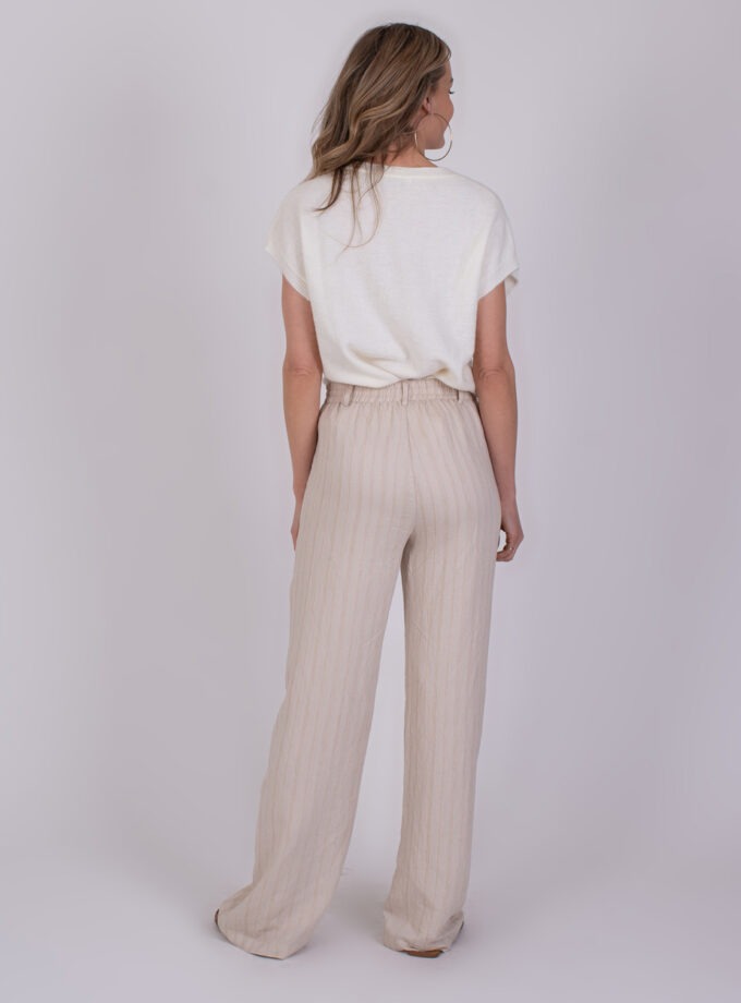 Vrouw gekleed in een witte top en beige, losvallende broek met strepen, gezien van achteren.