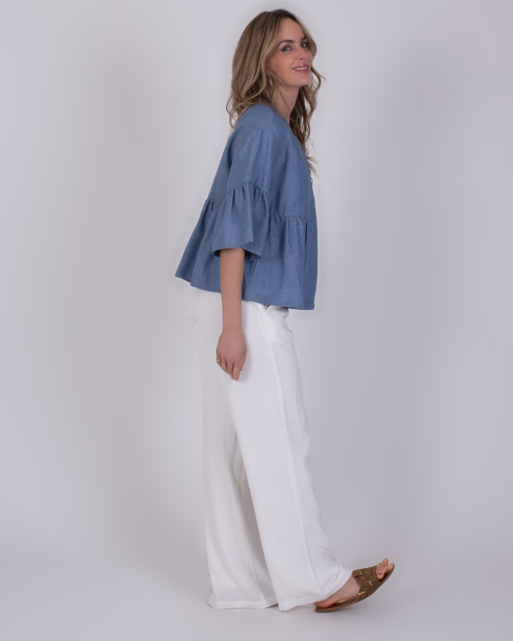 Khost light jeans blue 4 Vrouw in een Khost light jeans blue top met wijde mouwen en witte losse broek, gezien van opzij.