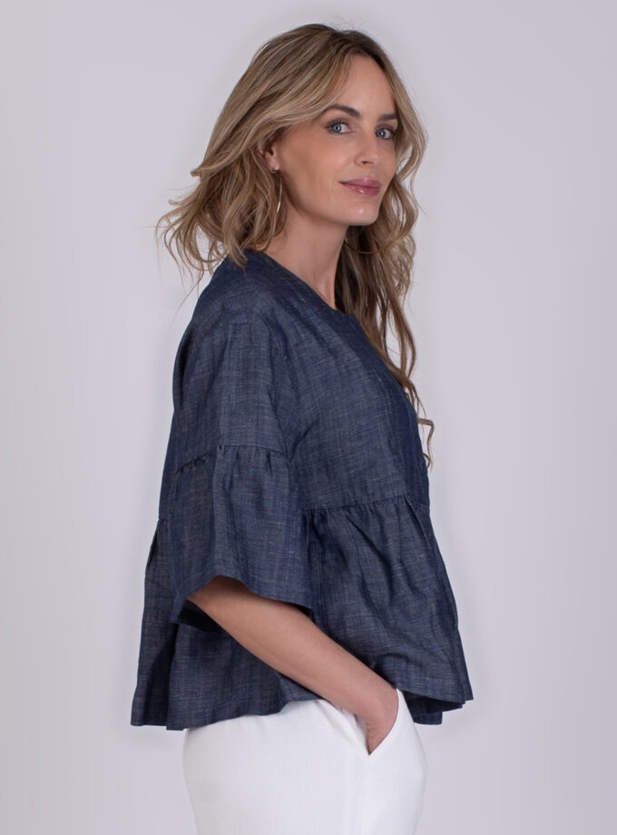 Vrouw in zijaanzicht draagt een donkerblauwe Khost-blouse met wijde mouwen en een witte broek.