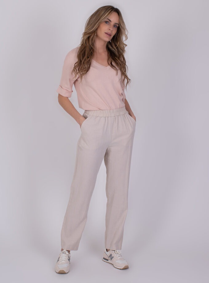 Vrouw draagt een lichtroze Korcula Pale 1-trui met beige broek en sneakers tegen een neutrale achtergrond.