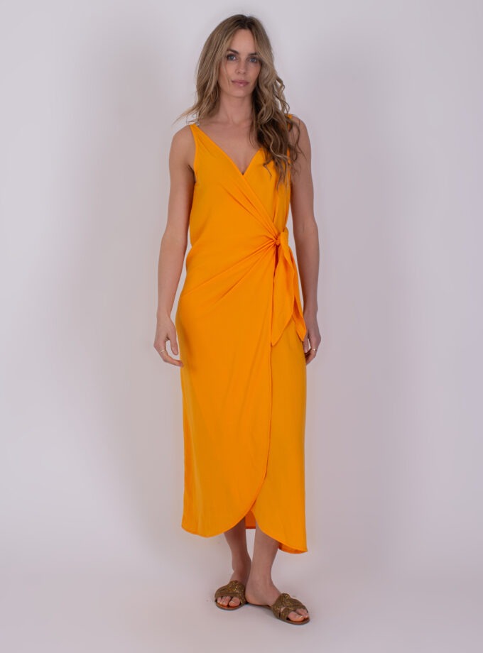 Vrouw draagt een mouwloze oranje Marlo-jurk met knoopdetail aan de zijkant.