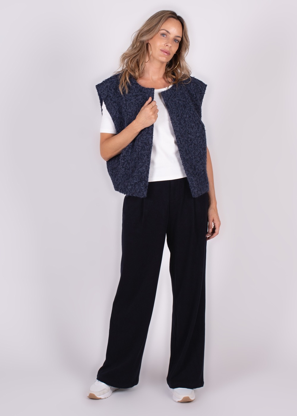 Een model draagt een donkerblauw, mouwloos Matsumoto gilet-70 over een wit T-shirt, gecombineerd met zwarte broek en witte sneakers, staande tegen een neutrale achtergrond.