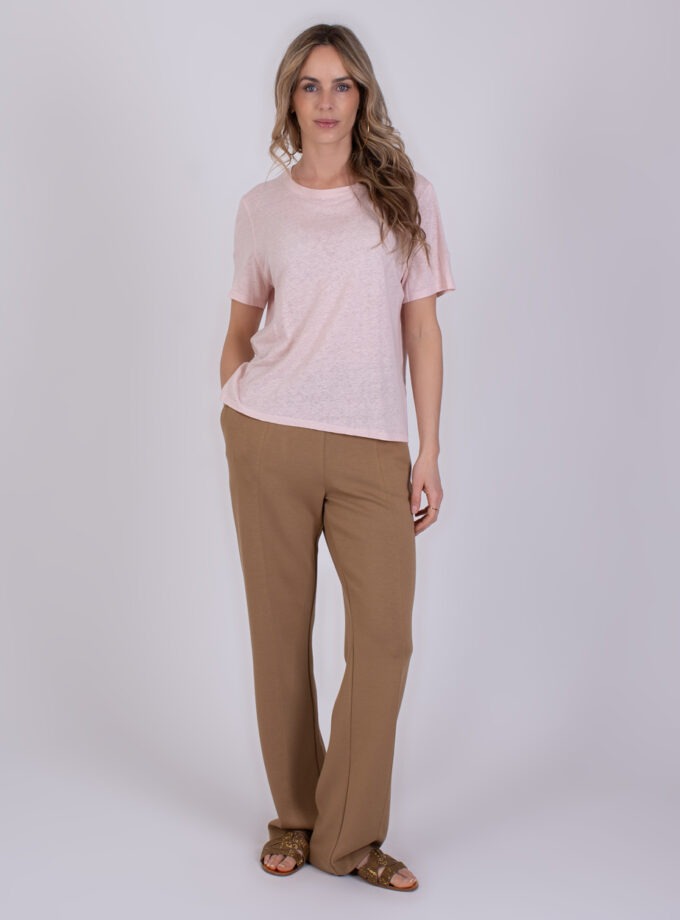 Vrouw draagt een lichtroze Monza T-shirt en beige broek met sandalen tegen een neutrale achtergrond.