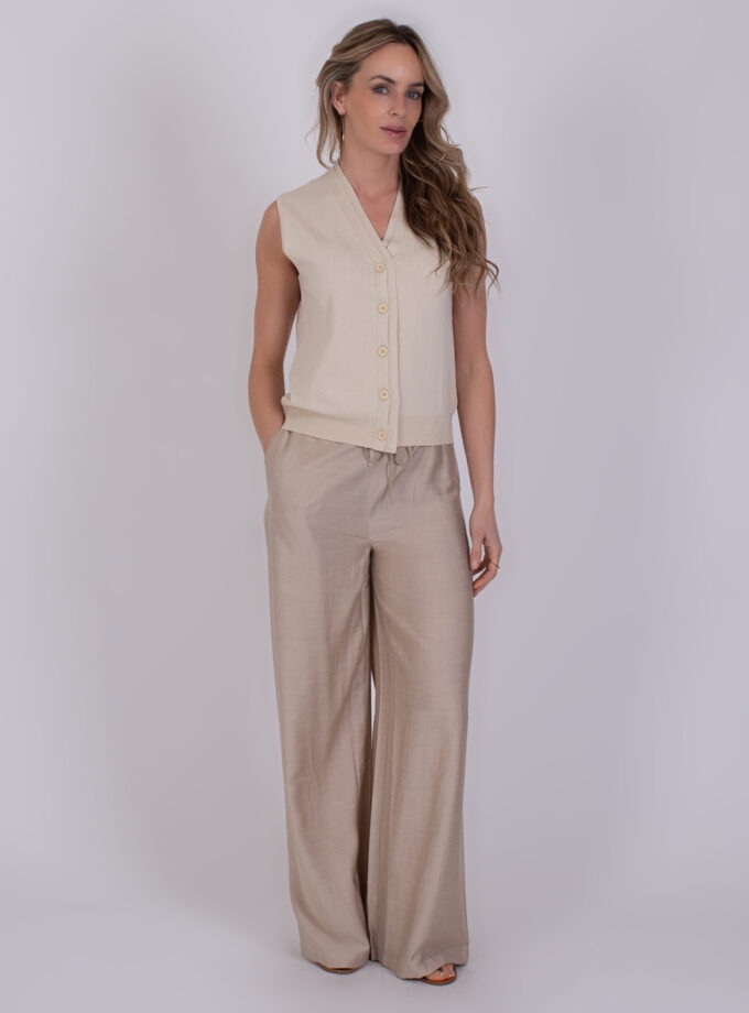 Vrouw draagt een mouwloos beige vest met knopen en wijde, lichtbruine broek uit de Patras Pale collectie.