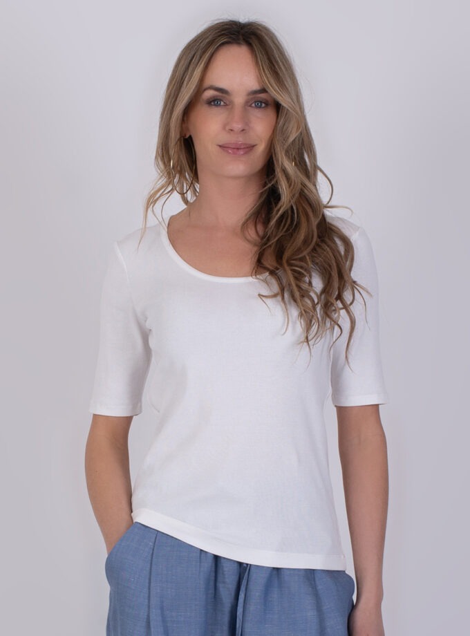 Vrouw draagt een offwhite T-shirt met halve mouwen en blauwe broek tegen een neutrale achtergrond.