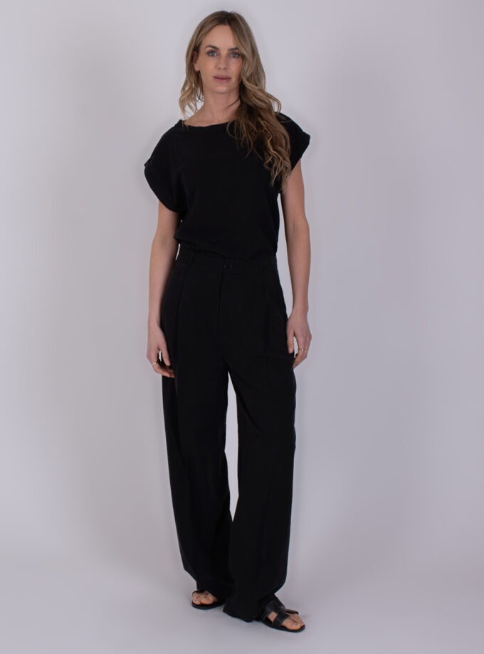 Vrouw draagt een zwarte Piran black 5 outfit met wijde broek en sandalen tegen een neutrale achtergrond.