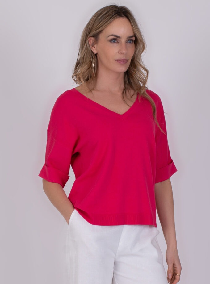 Vrouw draagt een fuchsiaroze Pylos-top met V-hals en opgerolde mouwen, gecombineerd met een witte broek.