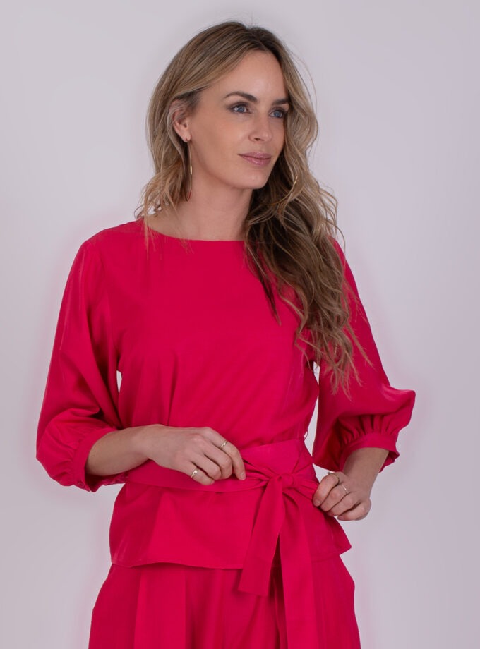 Vrouw met lang, golvend haar draagt een fuchsia Riyad-top met strikceintuur en bijpassende broek.