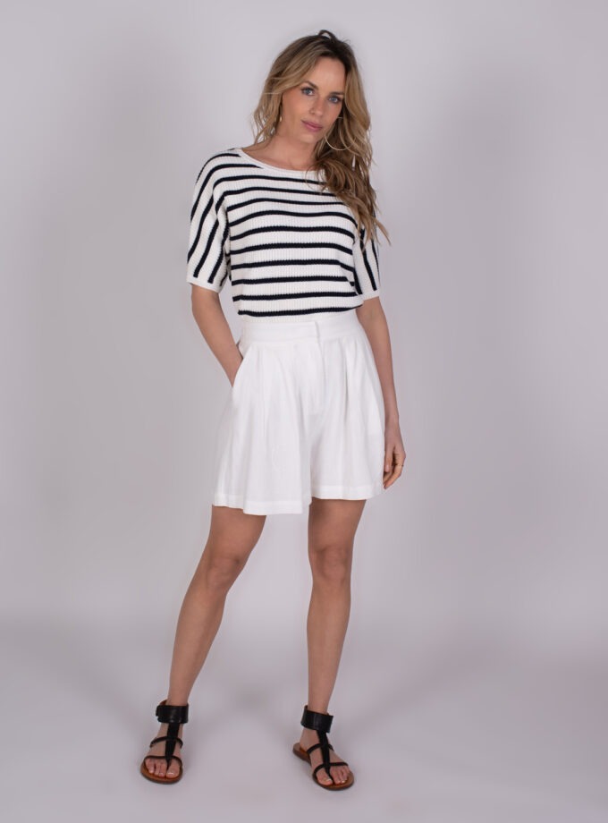 Vrouw poseert in een zwart-wit gestreepte trui met korte mouwen, witte shorts en zwarte sandalen tegen een neutrale achtergrond.