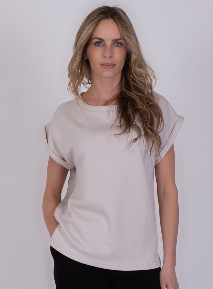 Vrouw draagt een beige Troje kit 1 T-shirt met opgerolde mouwen en zwarte broek.