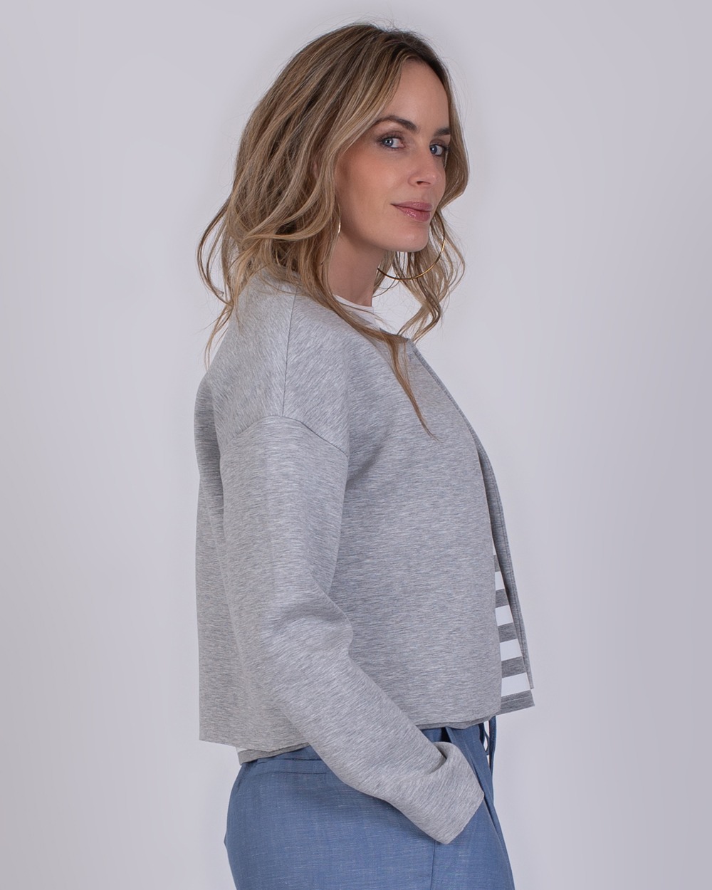 Tule grey 3 Vrouw draagt een grijs Tule vest met lange mouwen en een gestreepte binnenkant, gecombineerd met een blauwe broek.