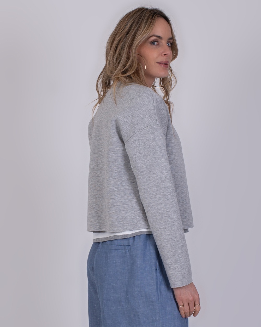 Tule grey 5 Vrouw in zijaanzicht draagt een grijze Tule Grey 5 trui en lichtblauwe broek tegen een neutrale achtergrond.