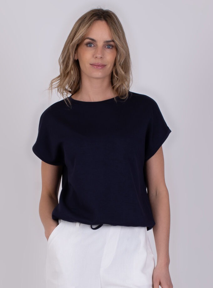 Vrouw draagt een donkerblauw Umea-shirt met korte mouwen en witte broek tegen een neutrale achtergrond.