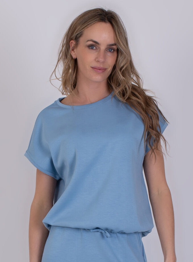 Vrouw draagt een lichtblauwe Umea-top met korte mouwen tegen een neutrale achtergrond.