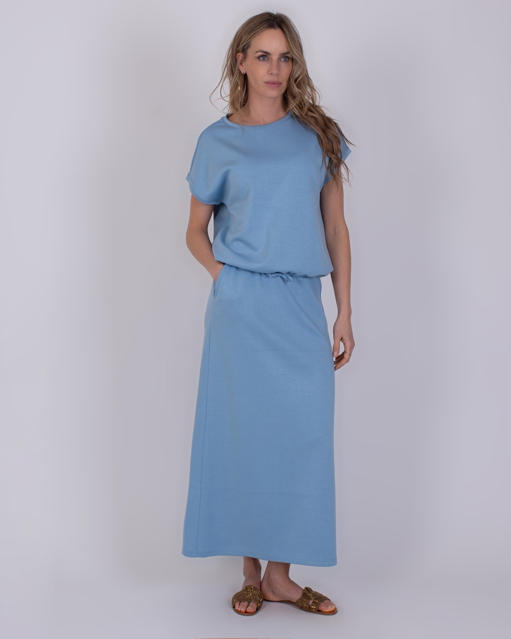 Umea soft blue 2 Vrouw draagt een lichtblauwe Umea-jurk met korte mouwen en een lange rechte rok.