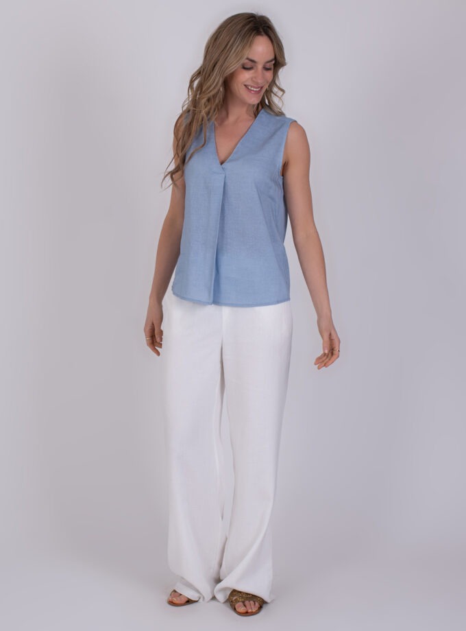 Vrouw draagt een mouwloze Valdris top in lichtblauw met een V-hals, gecombineerd met witte, wijde broek.