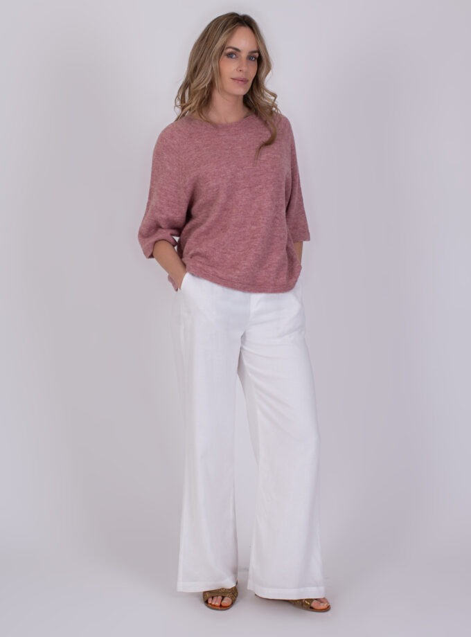 Vrouw draagt een losse, frambooskleurige Volos-trui met witte, wijde broek en sandalen.