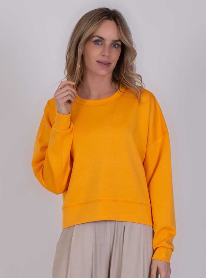 Vrouw draagt een feloranje York-sweater met een beige broek tegen een neutrale achtergrond.