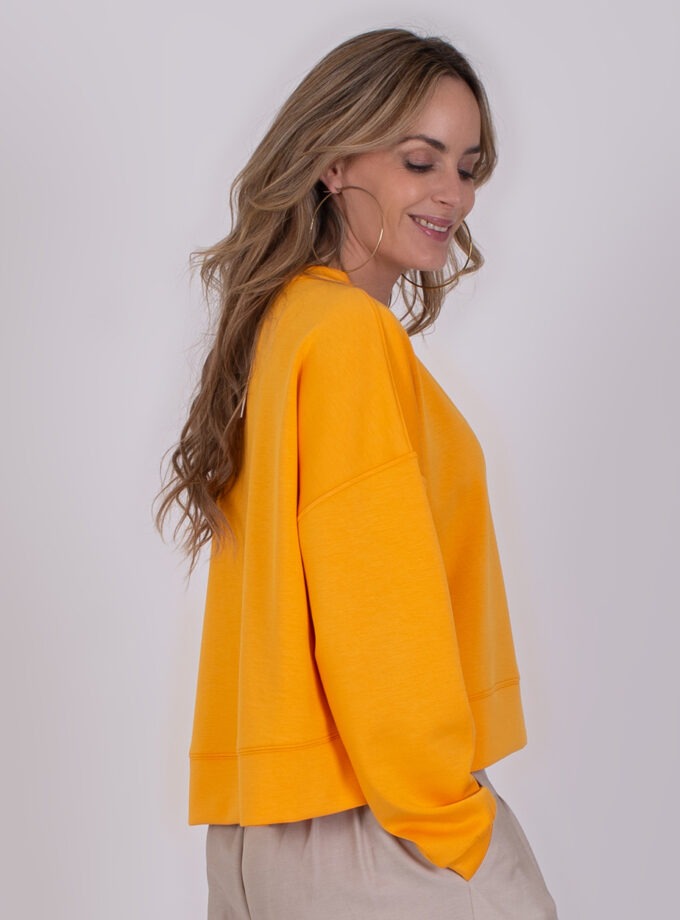 Vrouw in zijaanzicht draagt een feloranje York-sweater met beige broek.