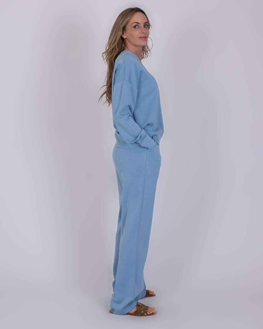 York soft blue 6 Vrouw in een comfortabele York soft blue loungetrainingspak, staand in zijaanzicht tegen een neutrale achtergrond.