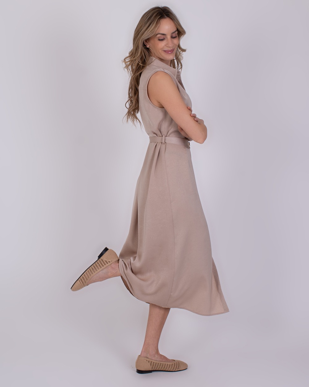 Zaria pale 5 Vrouw poseert zijwaarts in een mouwloze, beige Zaria Pale 5 midi-jurk met ceintuur en bijpassende schoenen.
