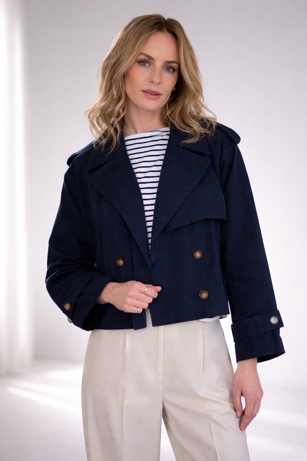 Een vrouw draagt een donkerblauwe, dubbelknoopsluiting jas genaamd "Aosta Navy" met een gestreept shirt en lichte broek.