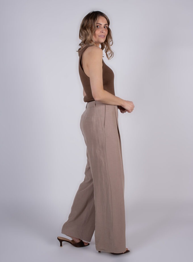 Een vrouw poseert zijwaarts in een bruine top met beige wijde broek en hakken voor een witte achtergrond.