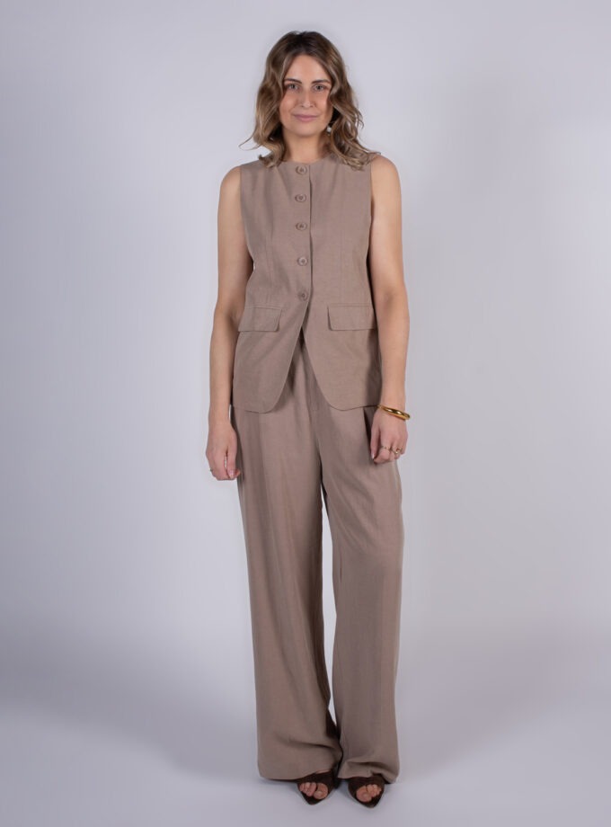 Een vrouw met halflang haar draagt een beige mouwloze blazer en broek, terwijl ze in een studio poseert.