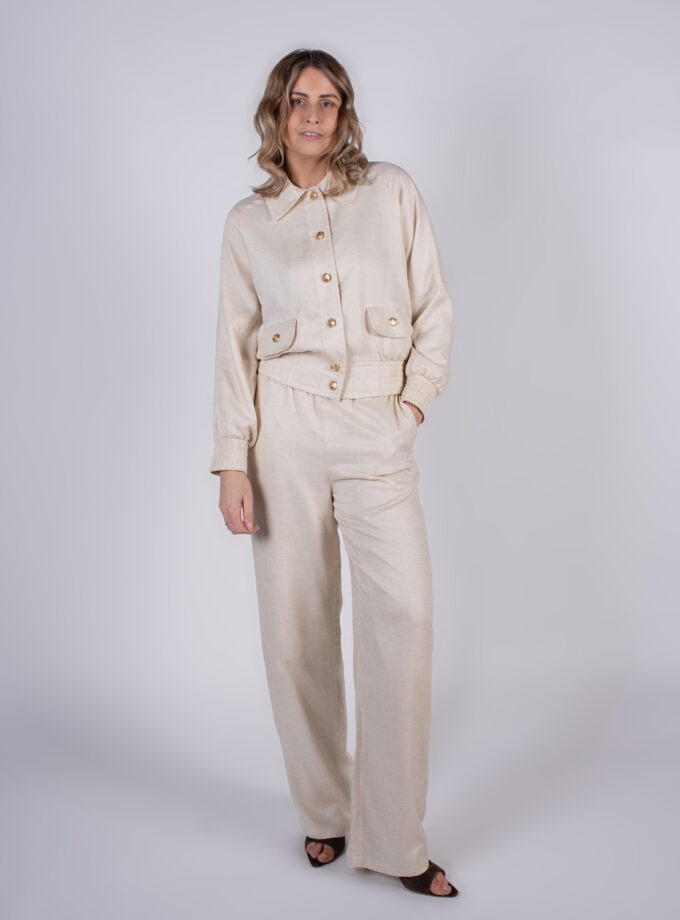 Model poseert in een beige, tweedelig ensemble met wijde broek en knoopjes op een witte achtergrond.