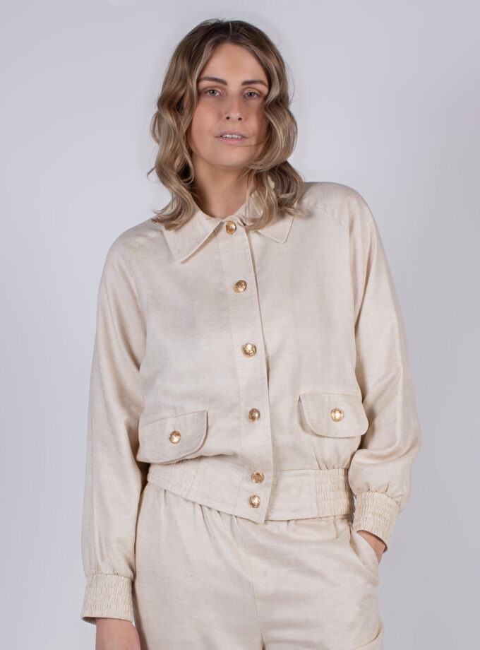 Een vrouw met los haar draagt een beige outfit met een jasje met goudkleurige knopen in een studio-opstelling.