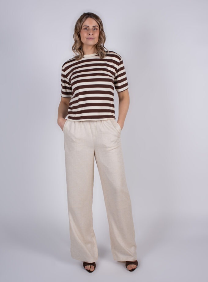 Een vrouw draagt een bruin-wit gestreept shirt en losse beige broek tegen een neutrale achtergrond.
