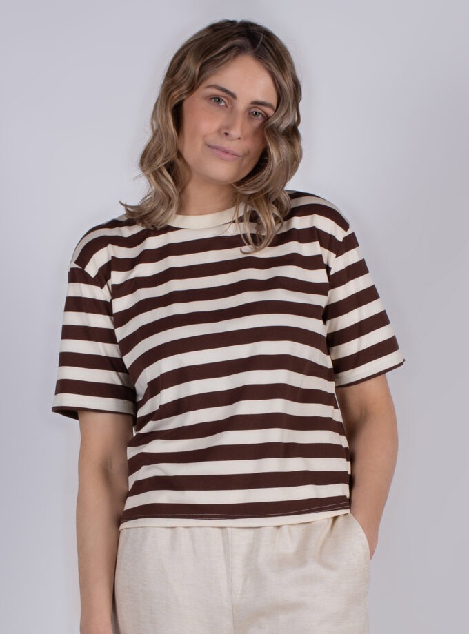 Een persoon met lichtbruin haar draagt een bruin met wit gestreepte T-shirt en beige broek tegen een grijze achtergrond.