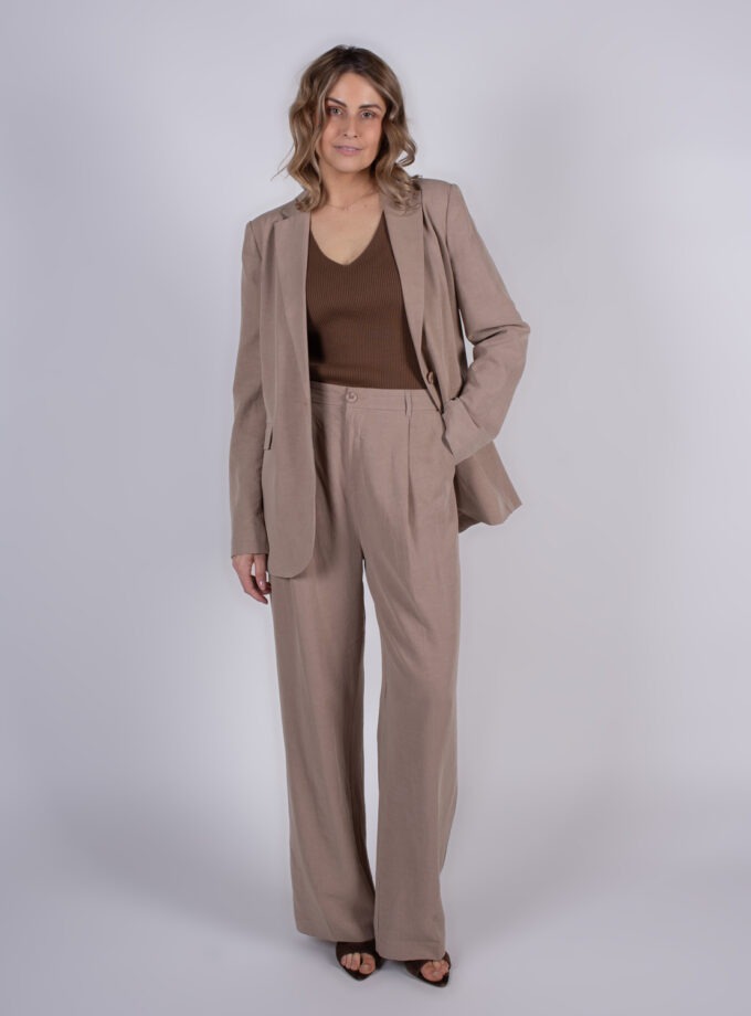 Een vrouw draagt een beige broekpak met een bruin bovenstuk en staat in een fotostudio.