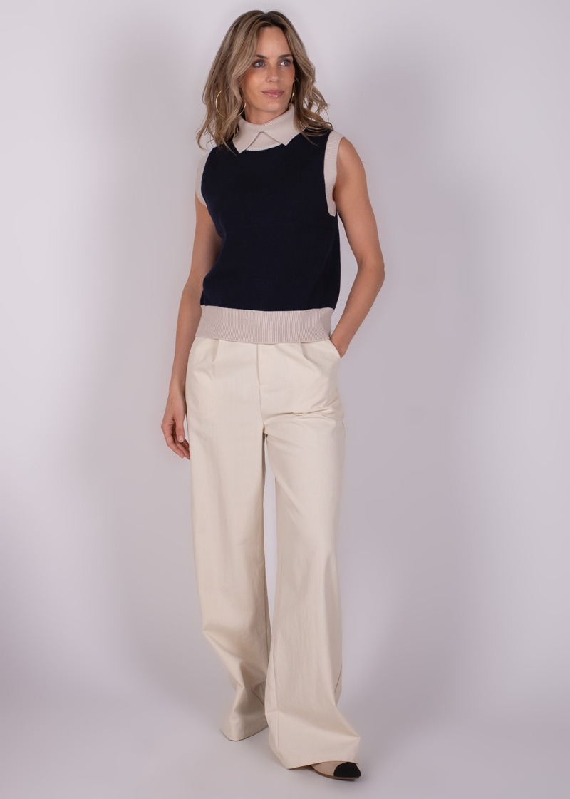 Model draagt een marineblauwe Arhus Spencer mouwloze trui met een beige boord over een beige blouse en wijde crèmekleurige broek, tegen een neutrale achtergrond.