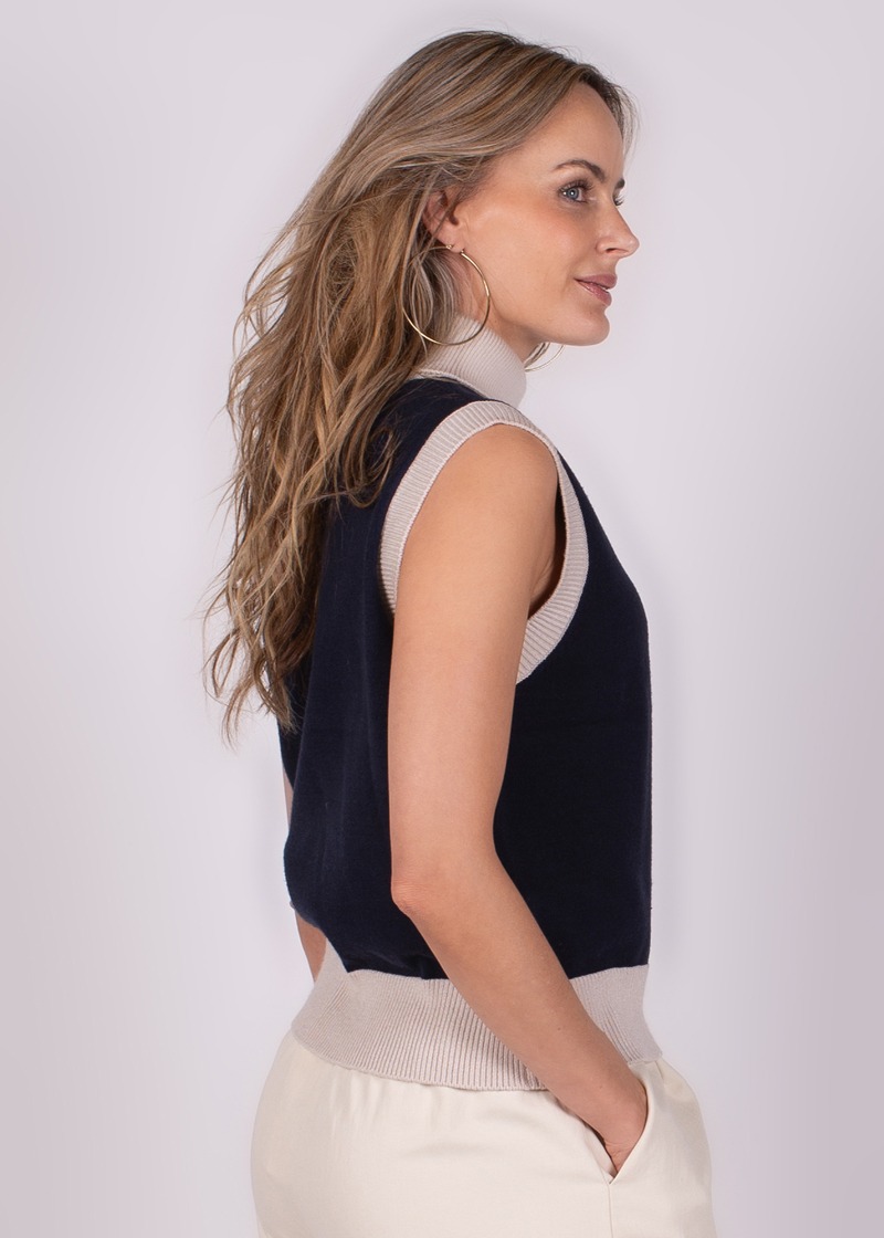Een vrouw in zijaanzicht draagt een marineblauwe Arhus Spencer zonder mouwen met beige boord, gecombineerd met een crèmekleurige broek.