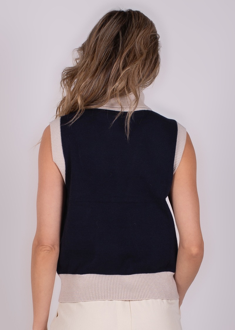 Een persoon met lang haar, gezien van achteren, draagt een donkerblauwe mouwloze Arhus Spencer met beige randen.