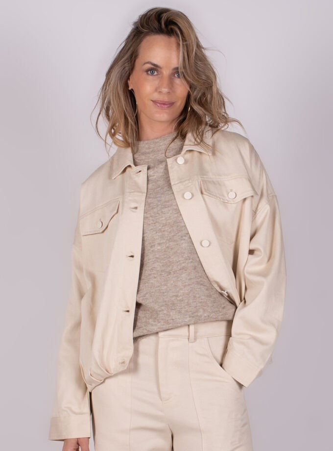 Een vrouw draagt een lichtgekleurde Balam jacket-124 met een beige trui en broek, gefotografeerd vanuit een frontaal standpunt.