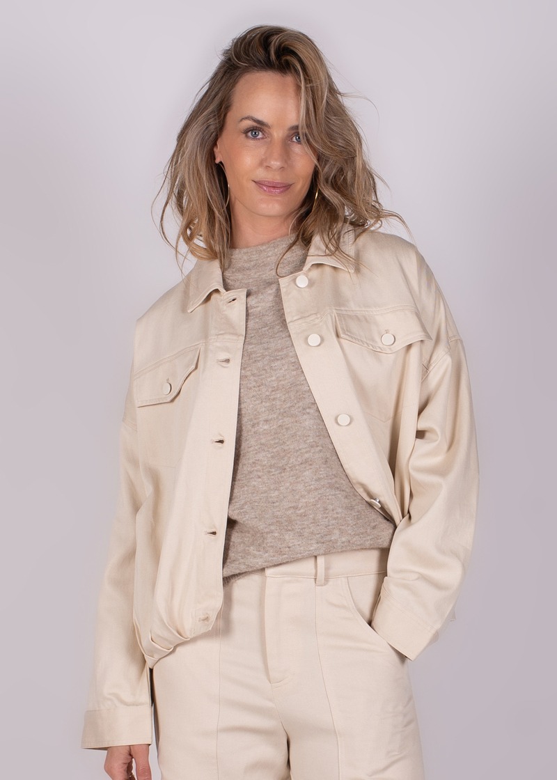 Een vrouw draagt een lichtgekleurde Balam jacket-124 met een beige trui en broek, gefotografeerd vanuit een frontaal standpunt.