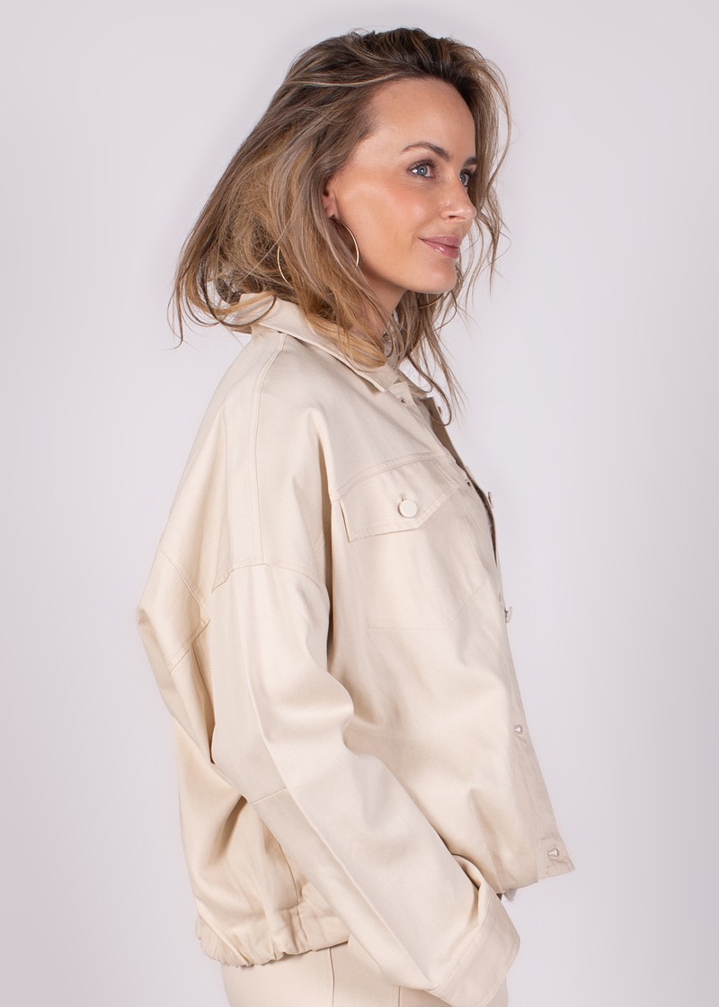 Vrouwelijk model poseert zijwaarts in een losse, beige Balam-jacket, met nonchalante blik naar rechts.