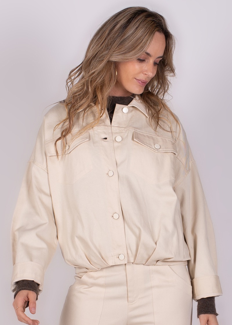 Een vrouw in een beige Balam-jack kijkt naar beneden, tegen een neutrale achtergrond.
