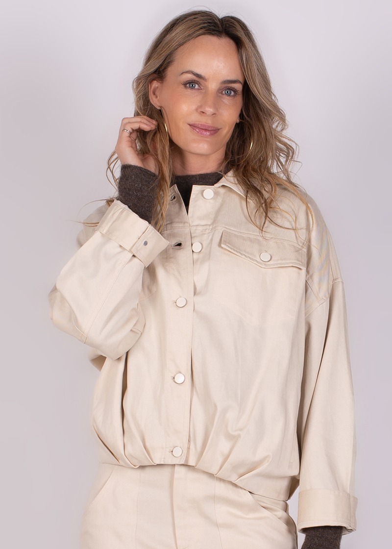 Vrouwelijk model met los haar draagt een crèmekleurige Balam jacket-135, kijkt naar de camera en houdt haar hand aan haar gezicht.