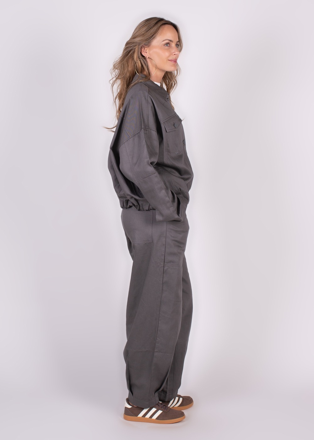 Een vrouw in een grijs kostuum genaamd "Balam pant grey 347" poseert zijwaarts tegen een witte achtergrond.