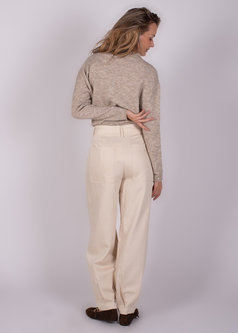Een vrouw gezien van achteren draagt een beige broek en een lichtbruine trui, terwijl ze naar links kijkt.
