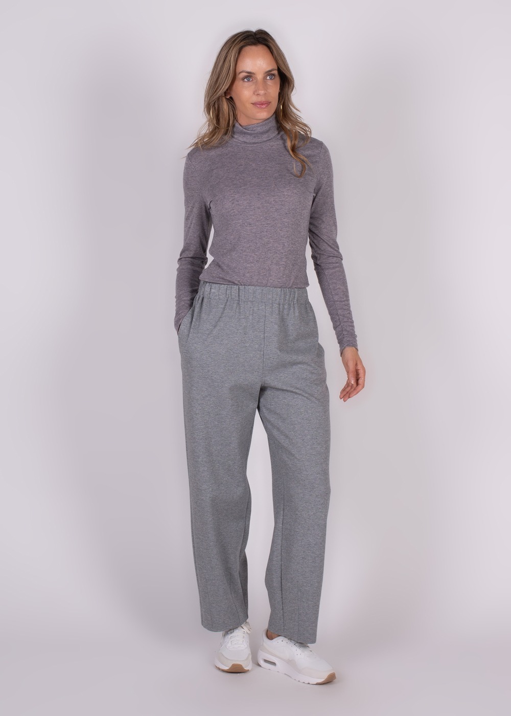 Een vrouw draagt een grijze Bilbao Barrel broek met een paarse coltrui en witte sneakers, poseert tegen een neutrale achtergrond.