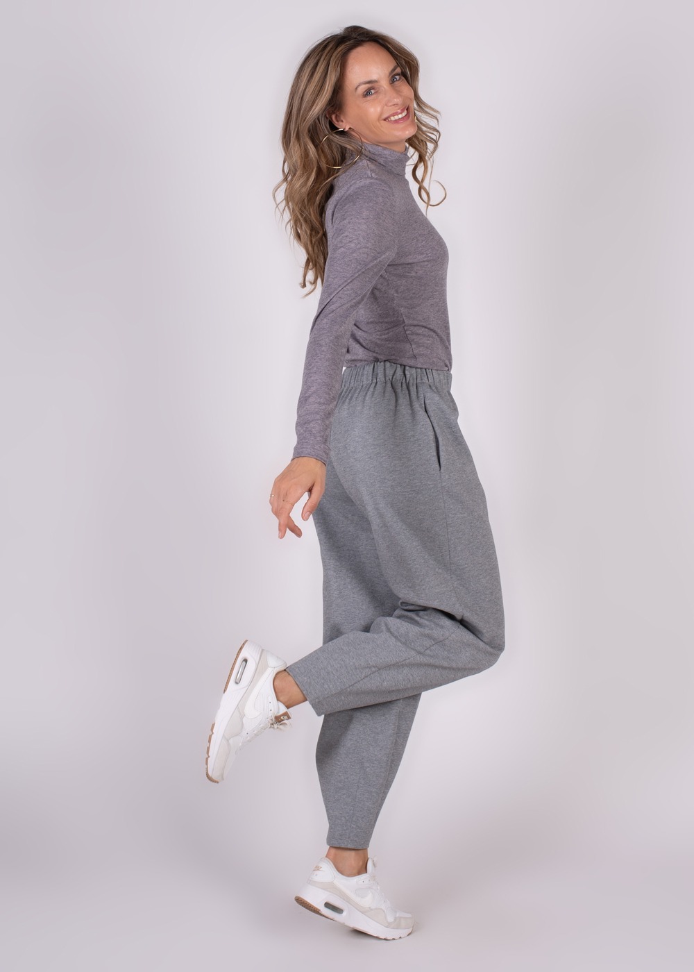 Een vrouw gekleed in een grijs T-shirt en Bilbao Barrel broek poseert speels zijwaarts, terwijl ze witte sneakers draagt.