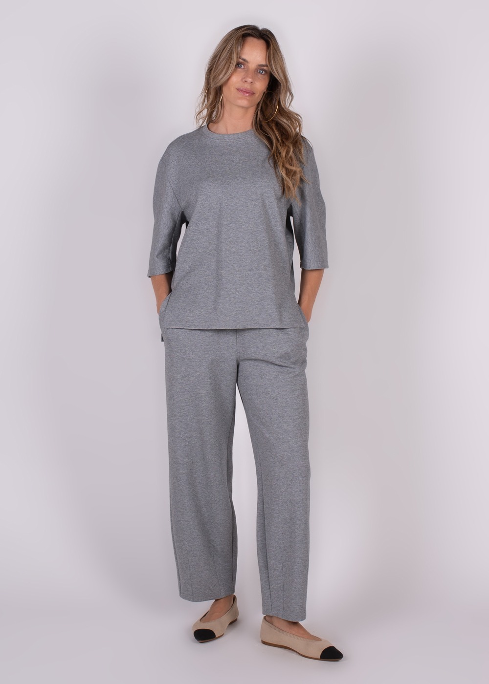 Model draagt een grijze outfit bestaande uit een top met driekwartsmouwen en wijde Bilbao-broek, gefotografeerd recht van voren.