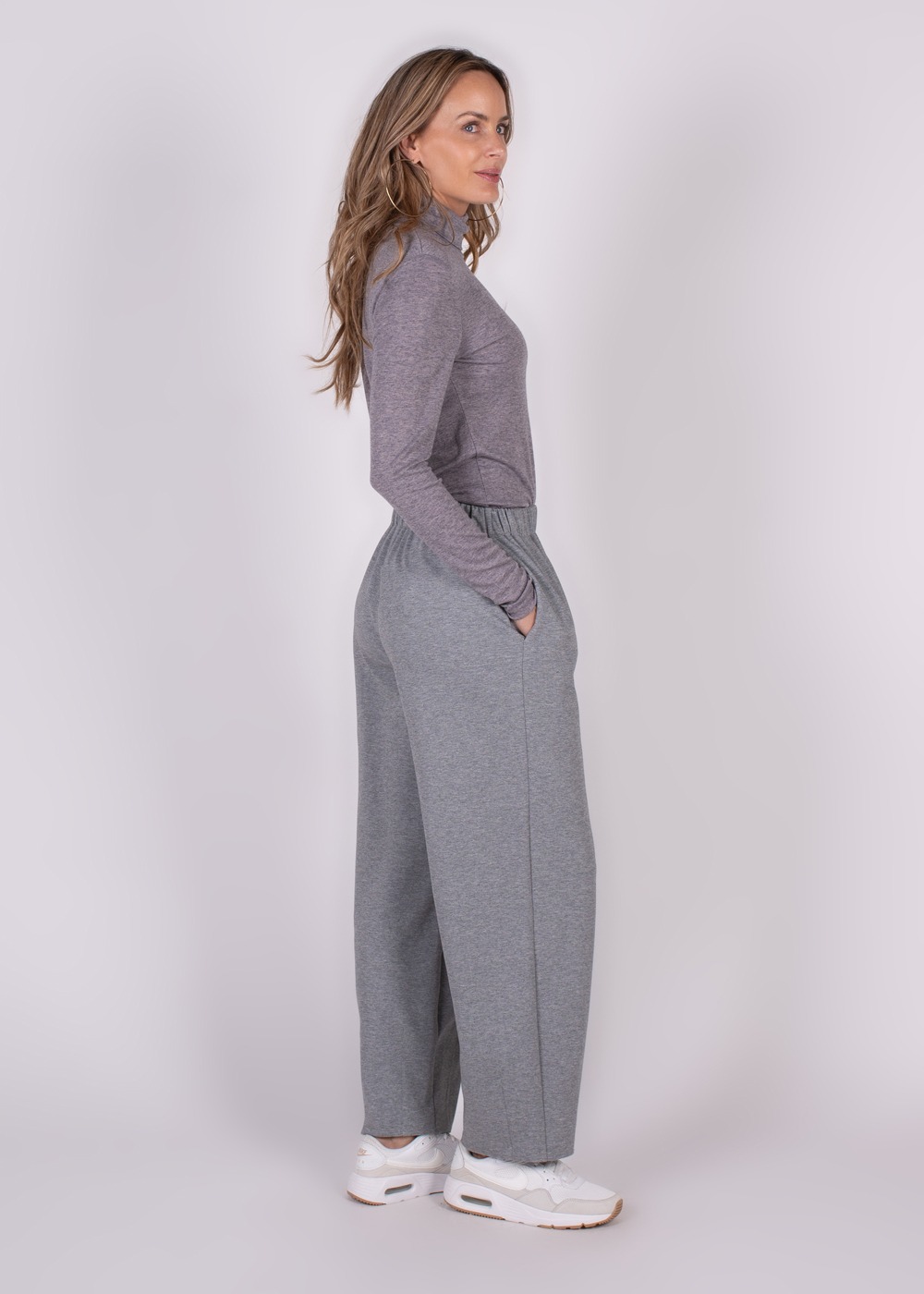 Model in zijaanzicht draagt een grijze Bilbao broek met elastische tailleband en witte sneakers.