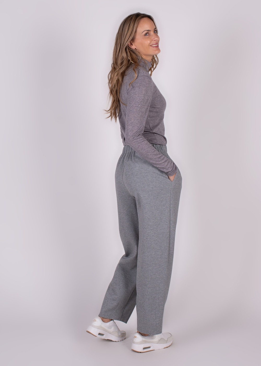 Een vrouw in zijaanzicht draagt grijze "Bilbao" broek en een grijze coltrui, met handen in de zakken en witte sneakers.