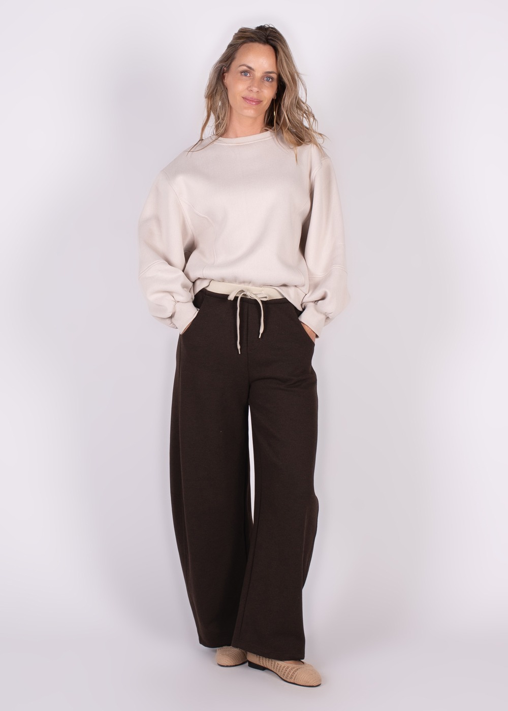 Model draagt een oversized Capri sweater in crème met bruine wijde broek en beige schoenen tegen een witte achtergrond.
