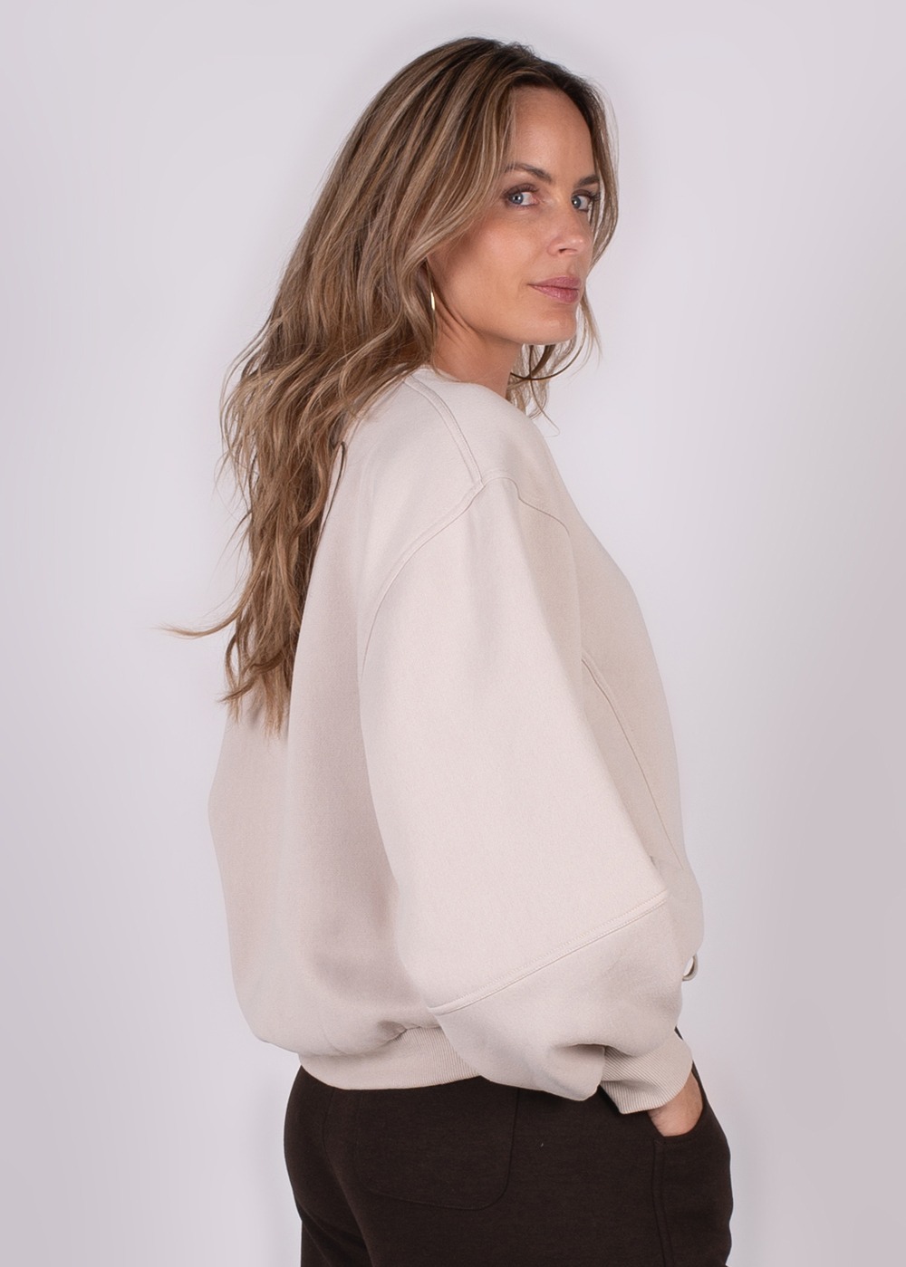 Model draagt een beige, oversized Capri sweater, gefotografeerd vanuit een zijaanzicht met haar los hangend.